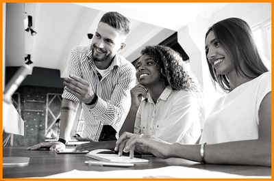 Three smiling coworkers collaborating on project looking at computer screen one typing two observing at table in office