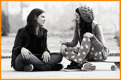 Two female friends sitting outside on pavement one with skateboard talking with empathetic character