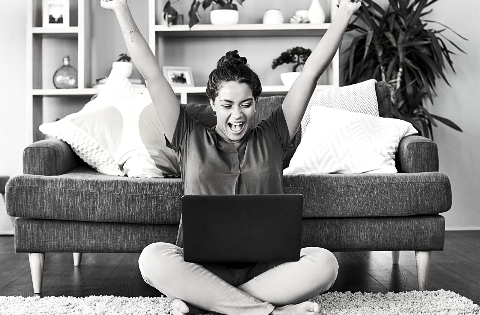 excited young woman sitting on living room floor with laptop arms in air showing she is enthusiastic person