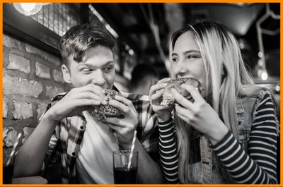 young couple at pub taking bites from sandwich looking into each others curious eyes