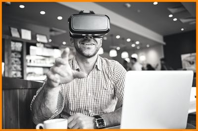 young curious professional man at coffee shop on laptop wearing virtual reality goggles smiling and reaching hand out