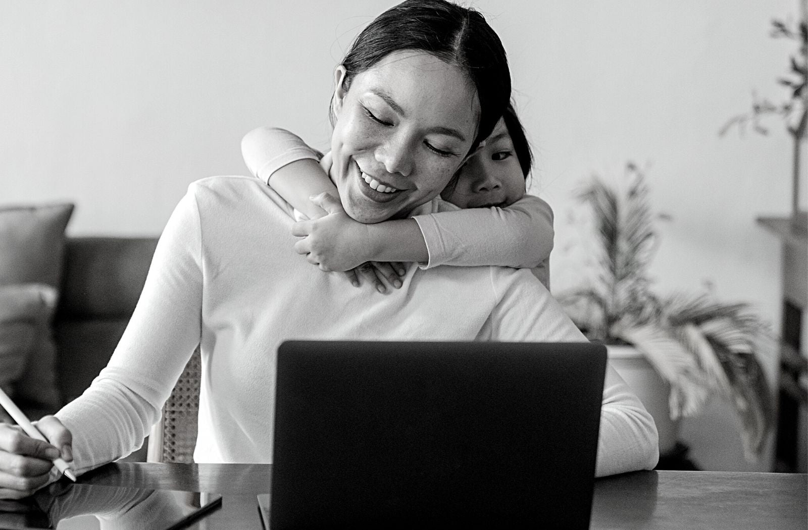 young mother with flexible character trait trying to work on laptop writing on tablet while daughter hugs her neck from behind